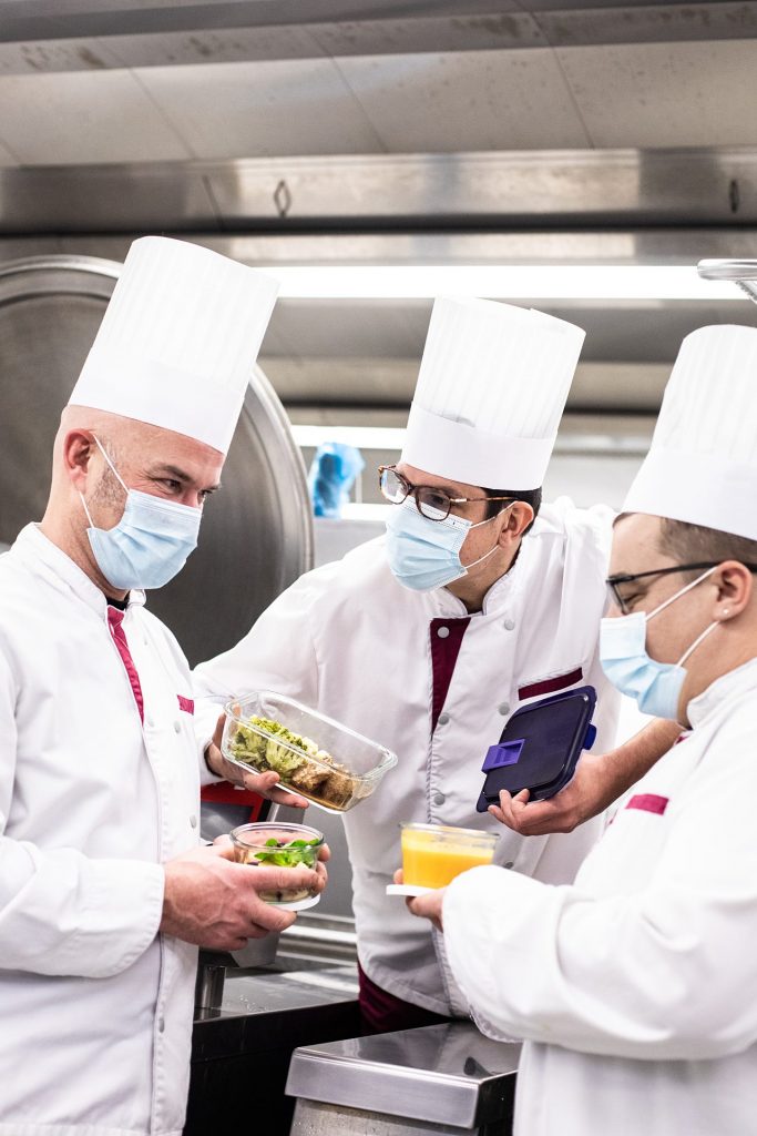 Trois chefs vêtus de blouses blanches et de masques, travaillant ensemble dans la cuisine de Tables Communes.